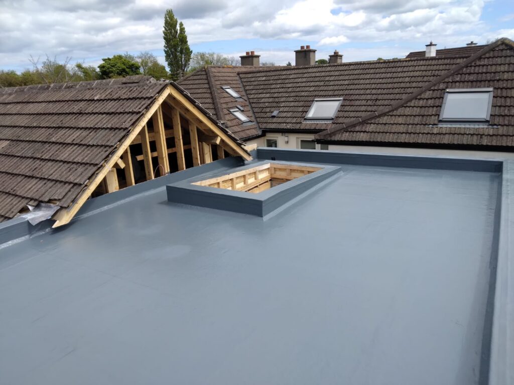 Overhead view of a completed grey GRP flat roof with a timber-framed rooflight upstand awaiting glazing, adjacent to an exposed dormer gable frame, with neighbouring concrete tile roofs featuring Velux windows visible in the background
