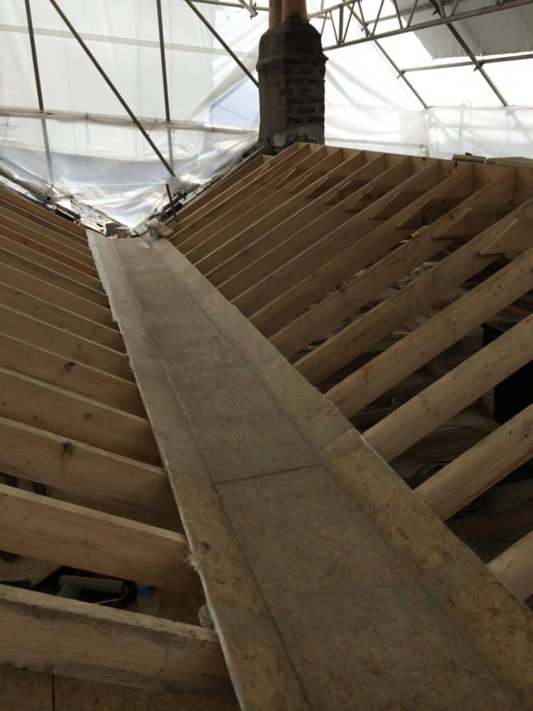 Looking up from inside a roof space showing exposed new timber rafters and ridge board forming a hip and valley junction, with a brick chimney stack visible and a temporary weatherproof scaffold cover overhead