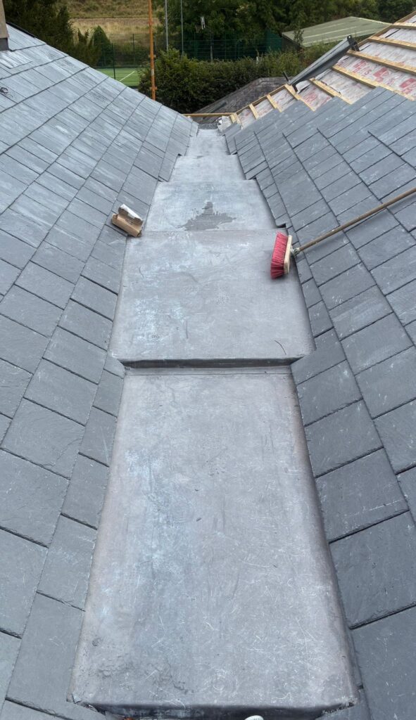 Partially completed natural slate roof showing a lead-flashed valley and stepped lead flashing detail, with a red-handled broom resting on the slates and an adjacent roof section still showing bare timber and breathable membrane