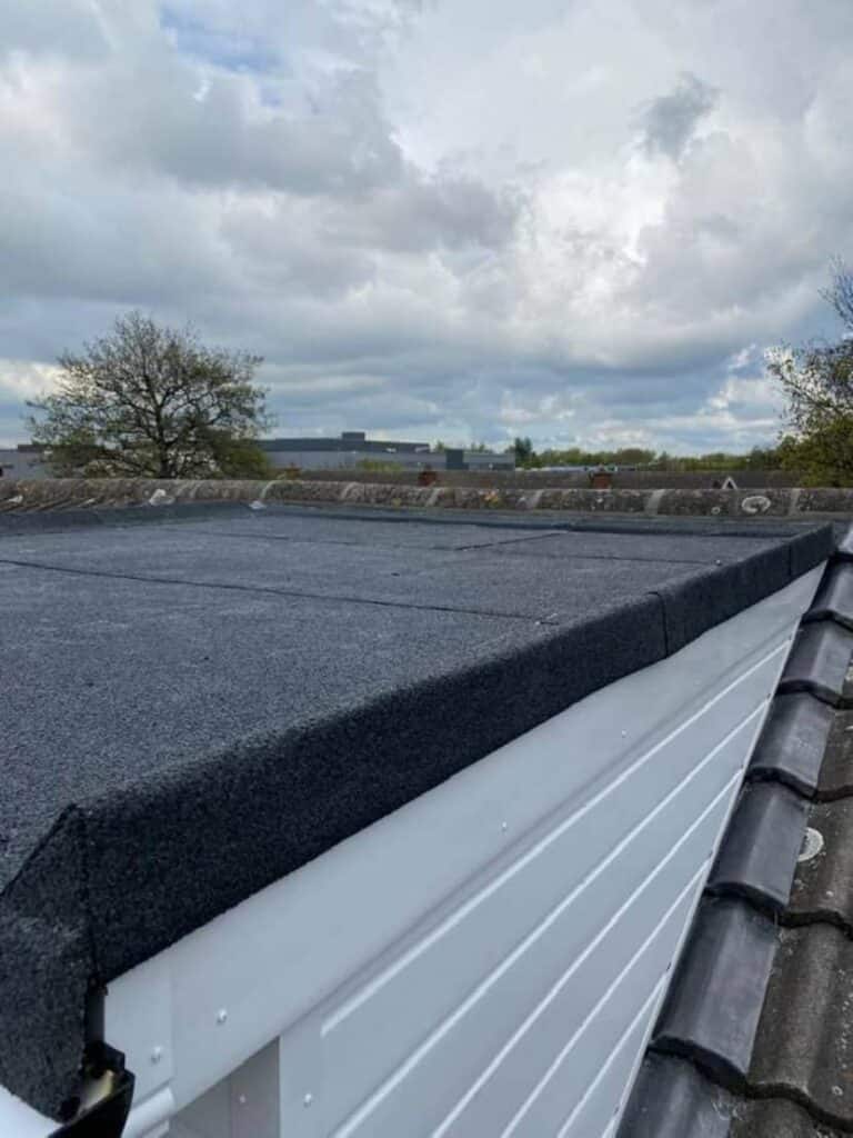 Close-up of a dark torch-on felt flat roof showing the finished edge wrapping over a white uPVC fascia board, with a concrete tile pitched roof meeting the flat section and open countryside and bare trees visible beyond the boundary wall