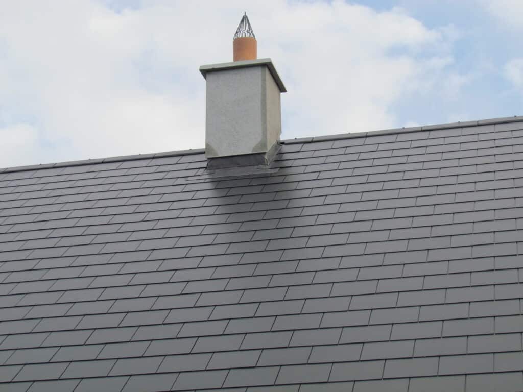 Close-up looking up at a large expanse of natural grey slate roofing with a rendered chimney stack featuring lead stepped flashing at the base, a terracotta chimney pot, and a metal bird guard at the top, against a partly cloudy blue sky