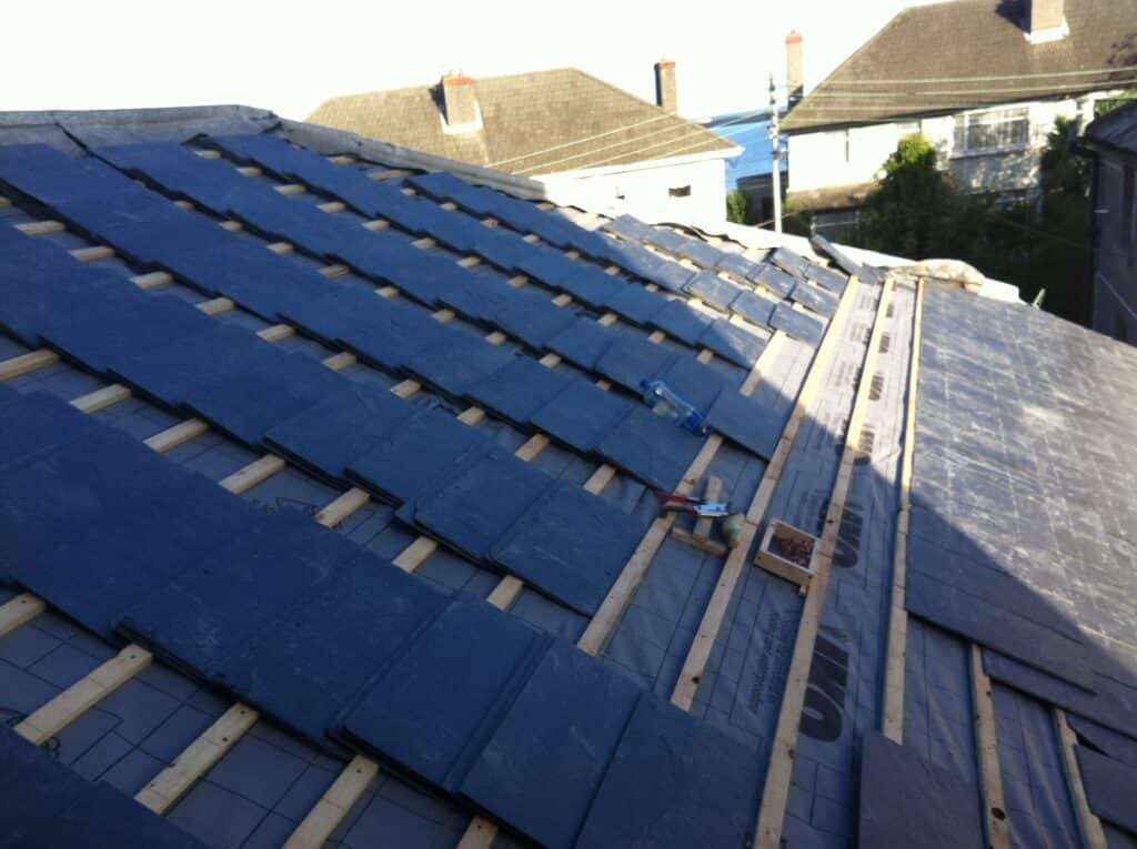 Close-up of a natural slate roof being laid over breathable underlay and timber battens, with a slate ripper and nails visible mid-course, and neighbouring rooftops in the background